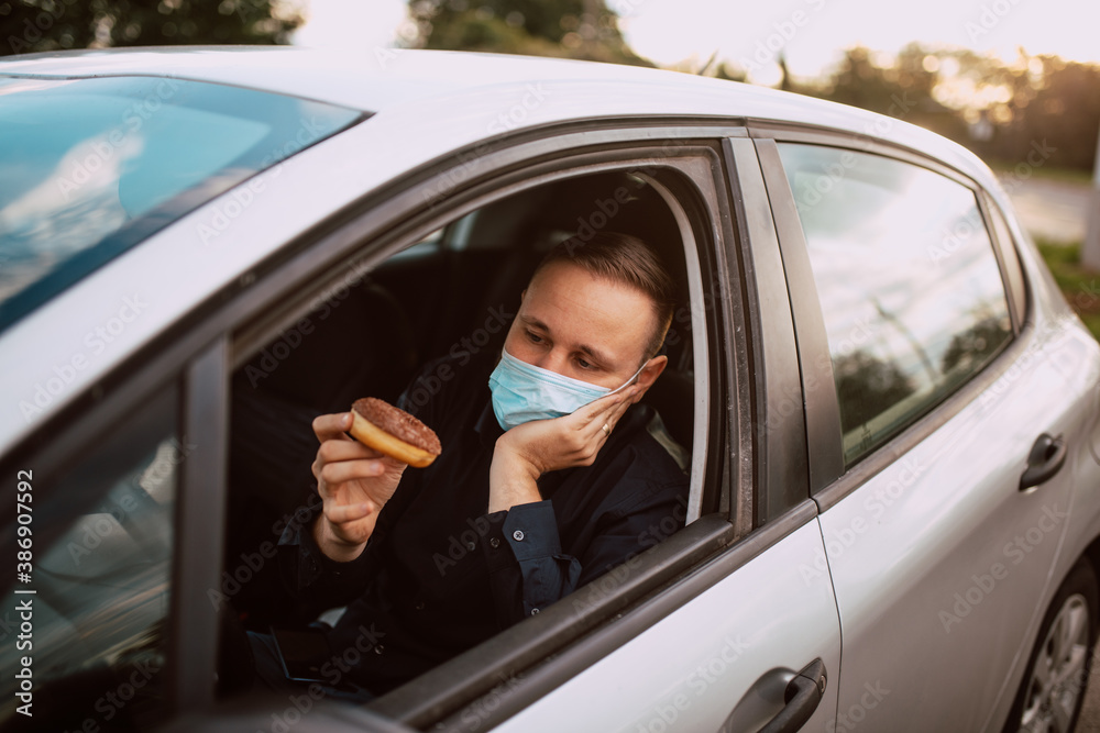 A portrait of a sad caucasian man with a medical face mask while sitting in a car while in traffic and holding a donut with chocolate. Meal in the car during the pandemic COVID - 19 coronavirus