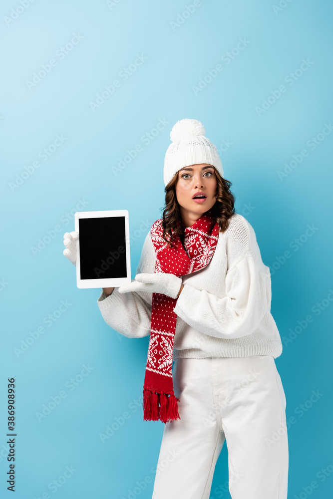 surprised young woman in winter outfit holding digital tablet with blank screen on blue