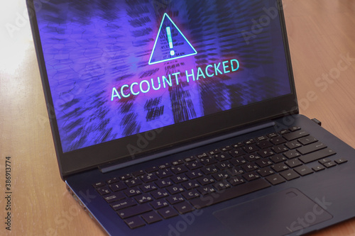 Laptop on the table and on a blue screen the text account is hacked. Warning triangular sign with exclamation mark symbol. Horizontal.