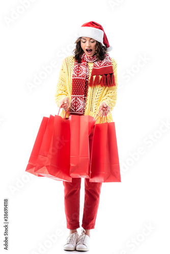 shocked young woman in santa hat and scarf with ornament holding red shopping bags isolated on white