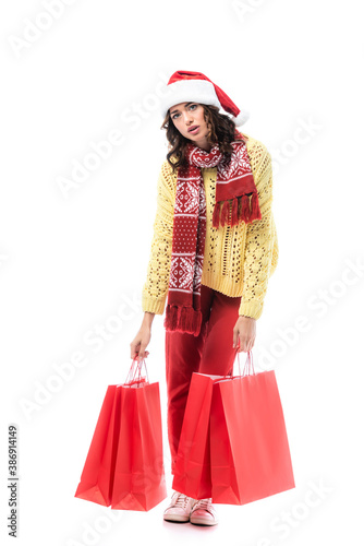 tired young woman in santa hat and scarf with ornament holding red shopping bags isolated on white
