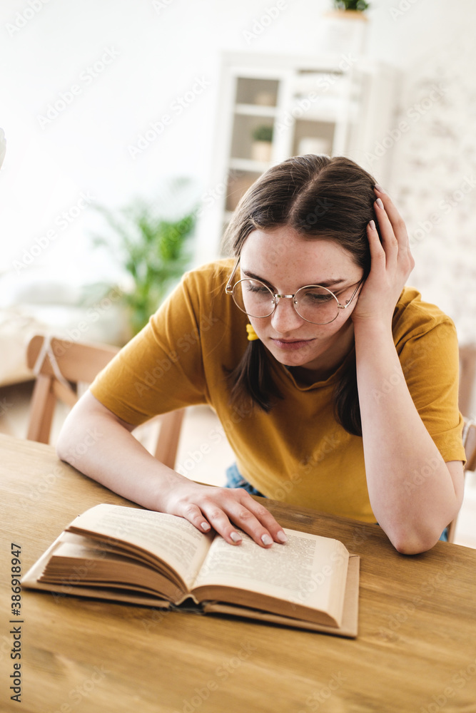 Young beautiful woman student in orange T-shirt and glasses reads book sitting at the table. Preparing for exams, studying at home on self-isolation. Young beautiful woman student in orange T-shirt and glasses reads book sitting at the table. Preparing for exams, studying at home on self-isolation.