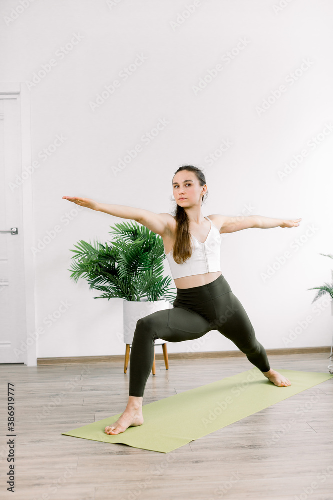 Healthy yoga and fitness concept. Young woman working out in cozy light room interior. Female yoga instructor doing warrior II, virabhadrasana two yoga pose on green mat indoors