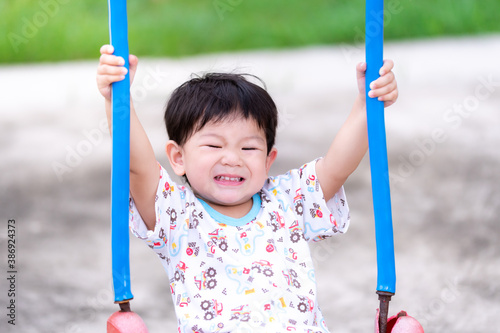 Boy plays on the swing, smiling sweetly. Toddler two hands caught on the blue string of the swings. Little children have fun in the playground. Child is 2 years old. © Kanthita