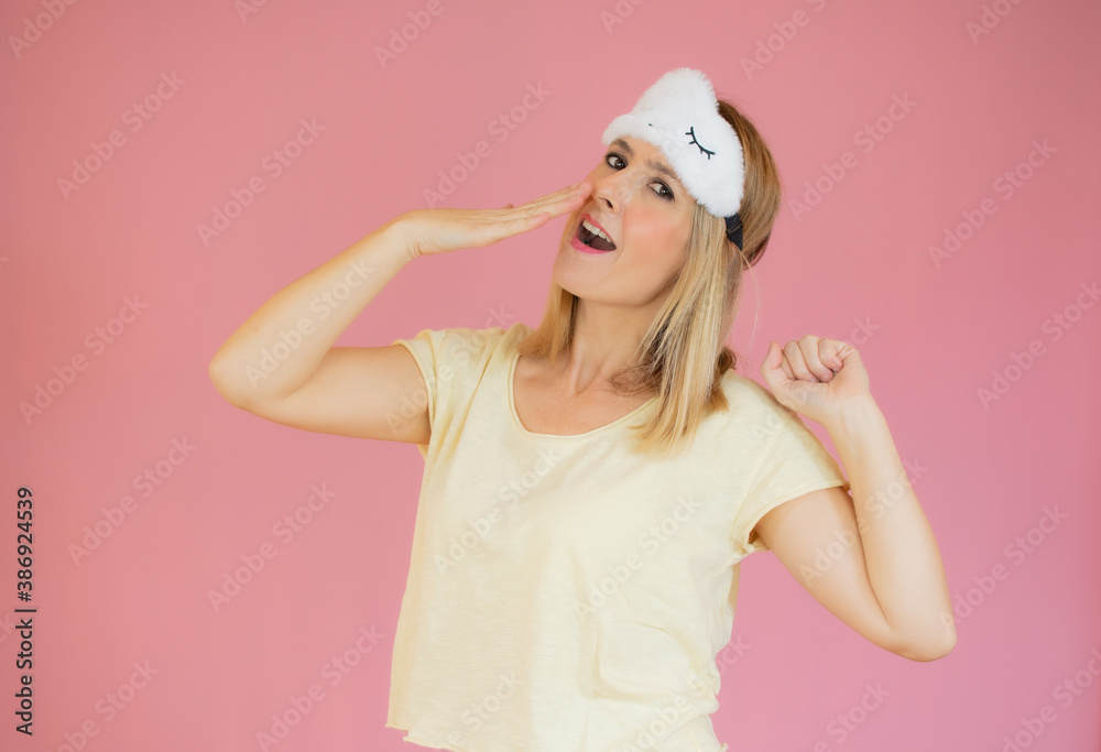 Tired young woman in yellow pajamas home wear sleep mask yawning covering mouth with hand resting at home isolated on pastel pink background studio portrait. Relax good mood lifestyle concept Tired young woman in yellow pajamas home wear sleep mask yawning covering mouth with hand resting at home isolated on pastel pink background studio portrait. Relax good mood lifestyle concept