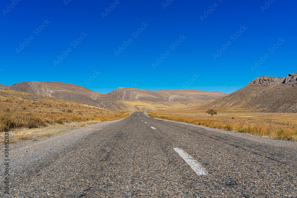 The road leads to the mountains. Mountain highway. Asphalt texture. Country highway. Mountains and hills in autumn. Mountain plants in autumn. Plants on the roadside. Dry yellow grass. Blue sky