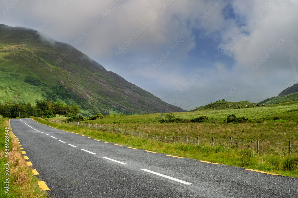 Naklejka premium Narrow empty asphalt road into mountains, Low cloudy sky over peaks. Nobody, Connemara, Ireland. Concept travel, road trip