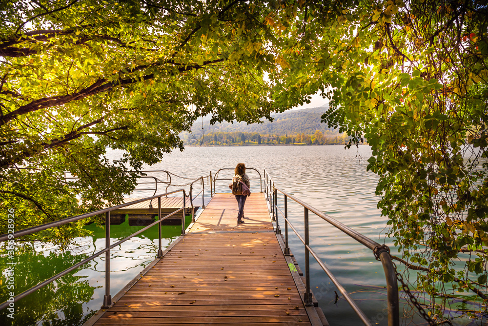Avigliana, Italy. October 10th, 2020. Woman walks on the jetty of the lake, framed through the branches showing autumn foliage.