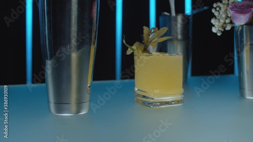 Close up shoot of the glass of Smash whiskey on the bar table. Rocks glass with ice, garnished with physalis and mint. Neon light tubes in the background. Motion control system.