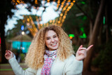 Portrait of Christmas happy smiling blonde curly woman wearing knitted white sweater and dancing or singing on decorated park background. Celebrating and festive mood. Soft selective focus.Copy space.