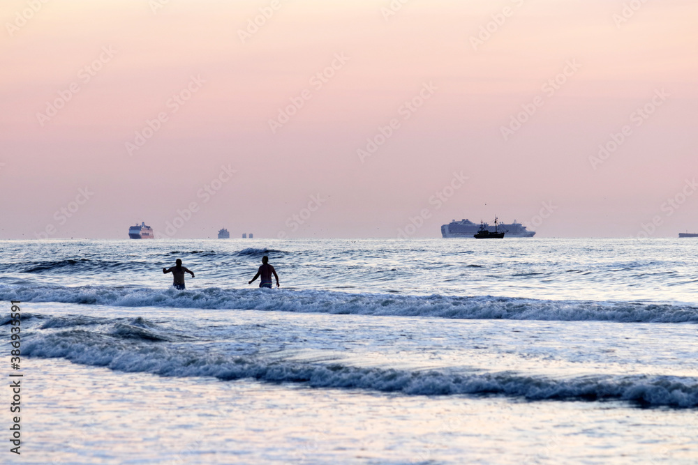 Naklejka premium Idle cruise ships parked near the Dutch beach where due to Covid pandemic at sunset; two person bathing in the water. 