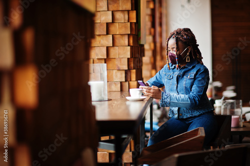 Stylish african american woman with dreadlocks afro hair, wear jeans jacket and face protect mask at restaurant, hold cellphone. New normal life after coronavirus epidemic.