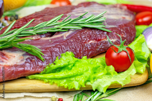 Raw red beef meat with green rosemary and fresh vegetables on light wooden cutting board background.