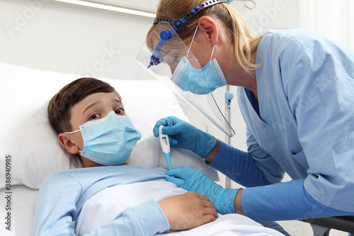 Nurse with thermometer measures the fever on patient child in hospital bed  wearing protective visor mask  corona virus covid 19 protection concept 