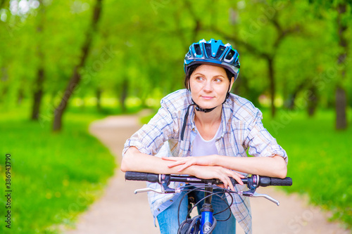 A girl in a bicycle helmet leans on the handlebars of a bicycle while walking in the park and looks thoughtfully aside