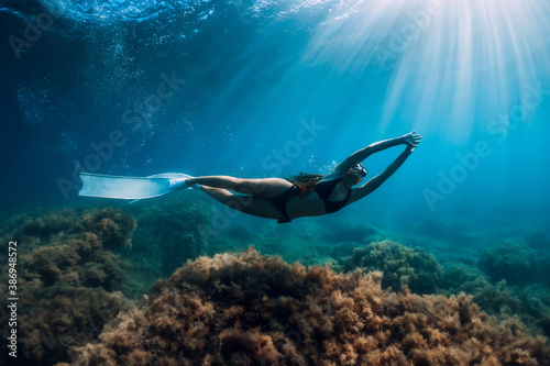 Freediver woman with white fins glides over seaweed bottom with sun rays. Freediving underwater in sea