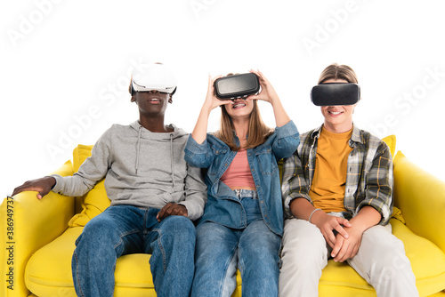 Smiling multicultural teenagers in vr headsets sitting on yellow couch isolated on white