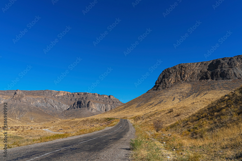 The road leads to the mountains. Mountain highway. Asphalt texture. Country highway. Mountains and hills in autumn. Mountain plants in autumn. Plants on the roadside. Dry yellow grass. Blue sky