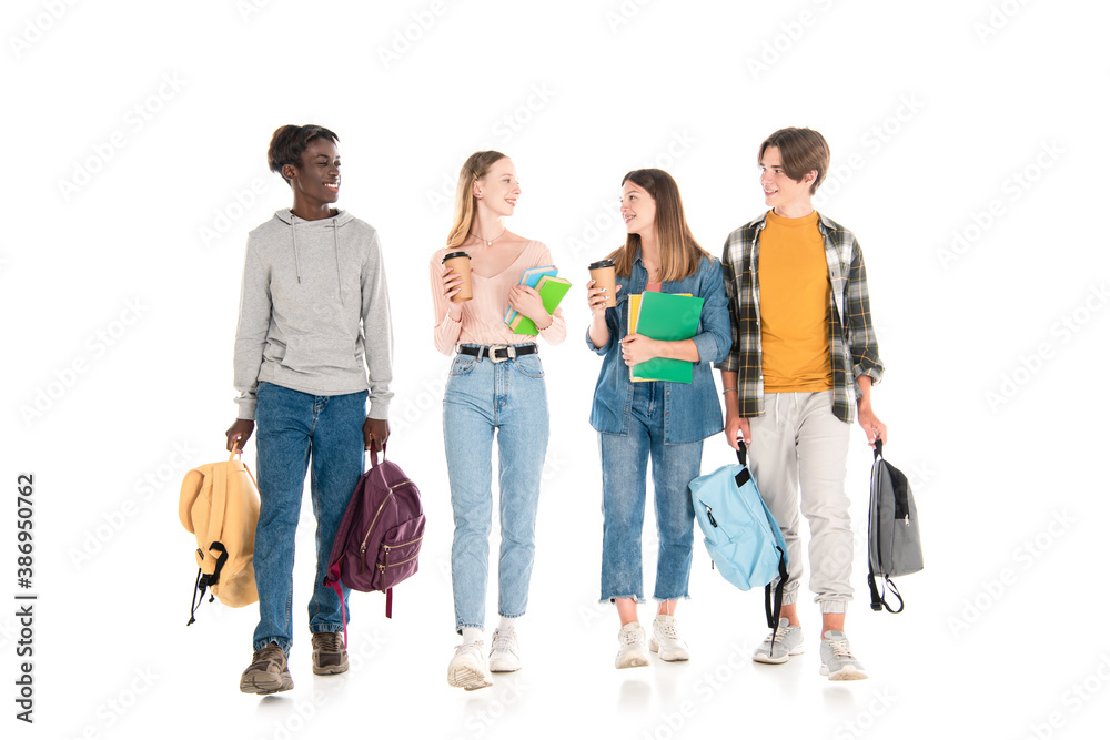Smiling multicultural teenagers with coffee to go, books and backpacks walking on white background