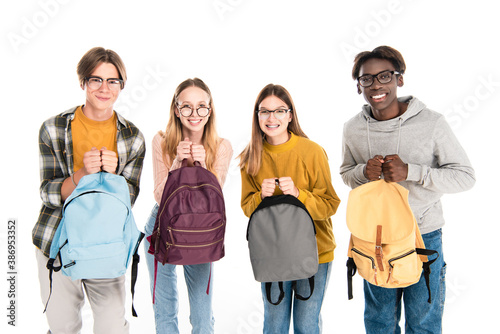 Smiling multicultural teenagers holding backpacks and looking at camera isolated on white