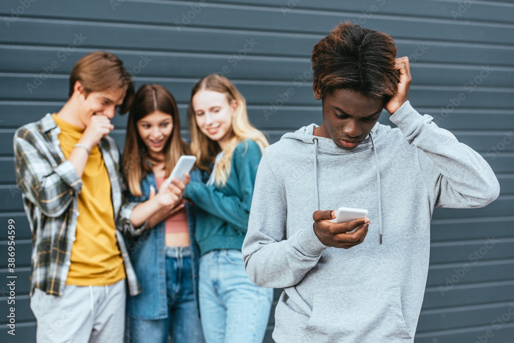 Fototapeta premium Selective focus of upset african american boy using smartphone near smiling teenagers with cellphone outdoors