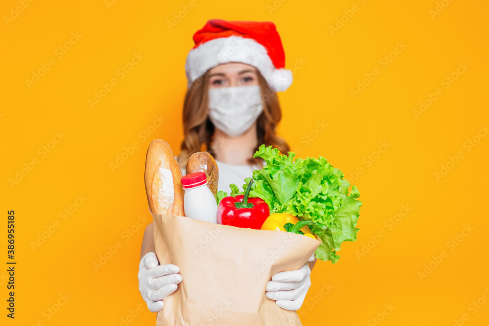 Safe food delivery for new year holidays. Courier girl in Santa Claus hat wearing a medical mask holds a paper bag with groceries, vegetables, herbs isolated on orange background Safe food delivery for new year holidays. Courier girl in Santa Claus hat wearing a medical mask holds a paper bag with groceries, vegetables, herbs isolated on orange background