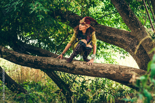 A wary and surprised elf sits on a tree branch and hides. Carnival and costume party. Magical forest. A woman dressed as an elv, she has elven ears and an elven hat.
