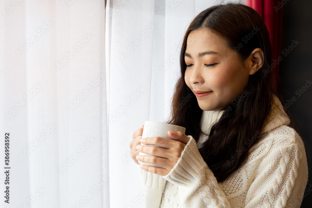 Young Asian woman in white sweater standing and holding coffee cup near window. Casual style indoor shoot Young Asian woman in white sweater standing and holding coffee cup near window. Casual style indoor shoot