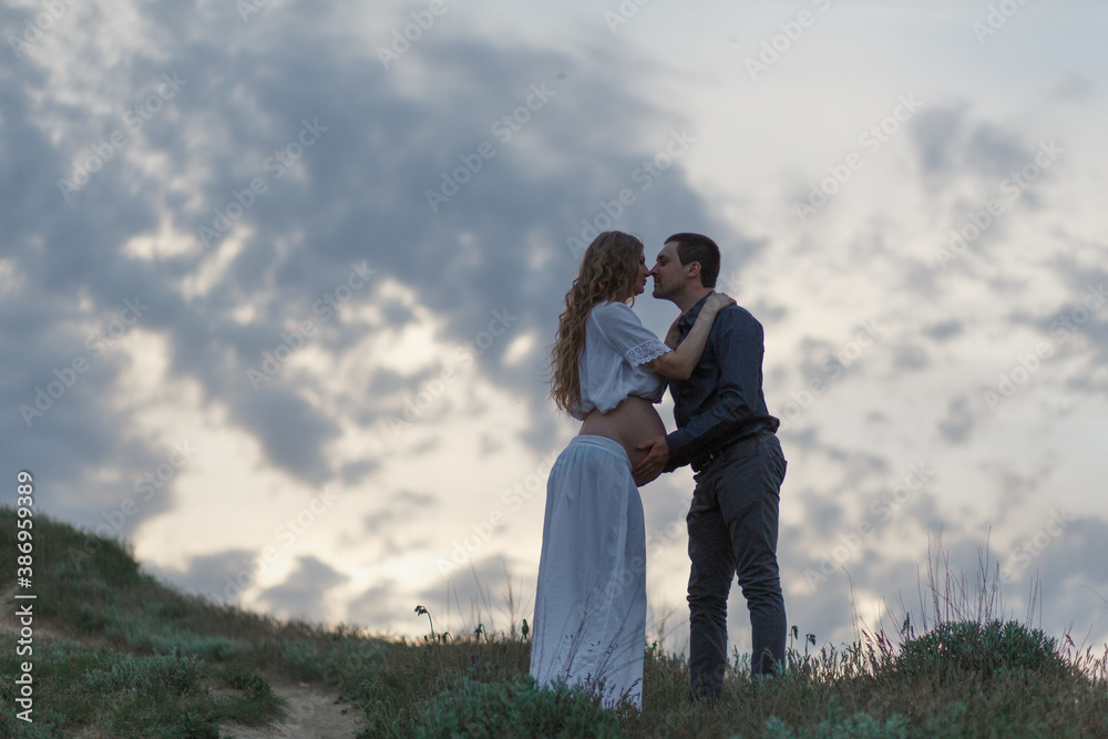 A couple in love, a pregnant wife in a white outfit are walking in a hilly field. Overcast weather A couple in love, a pregnant wife in a white outfit are walking in a hilly field. Overcast weather