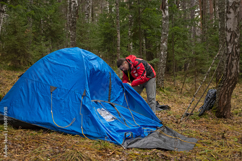 A young man in hiking clothes sets up a blue tent in the autumn forest. Tourism, activity, lifestyle.