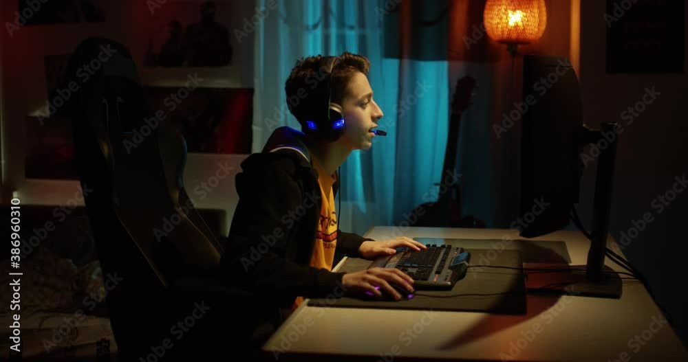 A teenage boy in headphones playing an online computer game, communicating with players, happy and excited