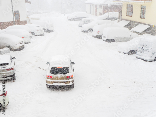 The car drives through a snow-covered yard in a blizzard past parked cars covered with snow
