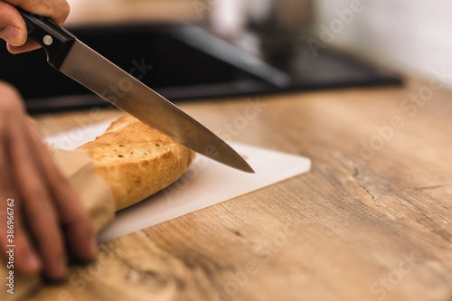 Crusty baguette on wooden cutting board with knife and man's fingers.