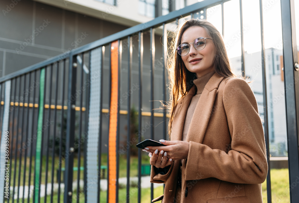 Fototapeta premium Smiling curly businesswoman wearing trendy sunglasses walks down the central city street and uses her phone. Businesswoman holding cell phone.