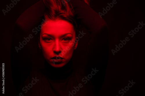 Low key portrait of pretty full-face girl raised long hair with her hands looking at camera in red twilight copy space. Attractive fashion model posing on dark studio background