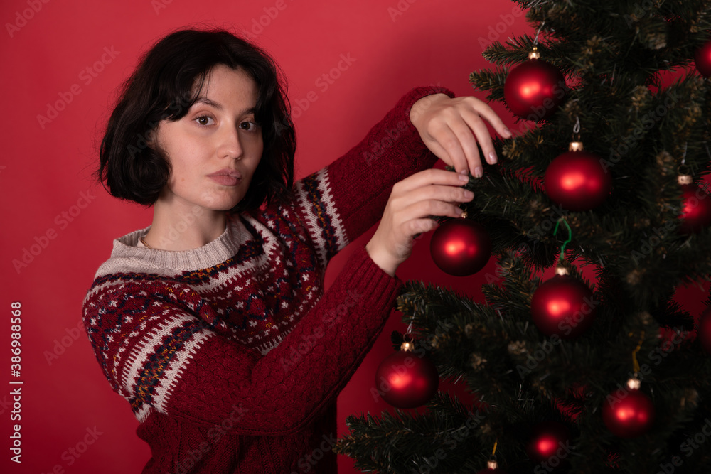Brunette girl dressed in knitted winter pattern sweater hanging toy balls on Christmas tree on studio red backdrop. Preparing celebrate New Year in quarantine alone. self-gifter concept. Brunette girl dressed in knitted winter pattern sweater hanging toy balls on Christmas tree on studio red backdrop. Preparing celebrate New Year in quarantine alone. self-gifter concept.