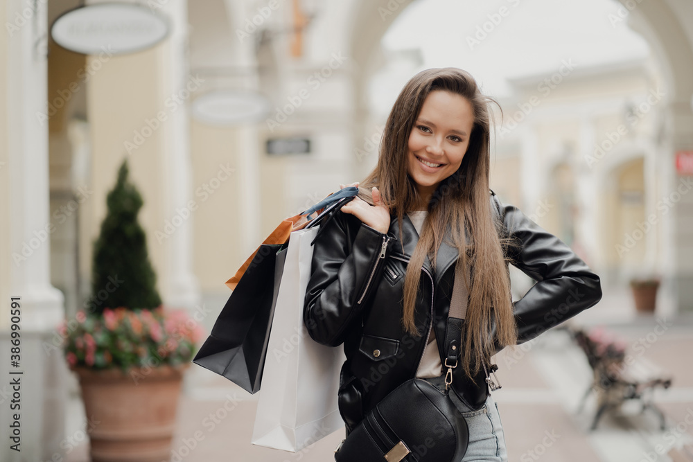 a brunette of European appearance in a black leather jacket and blue smiles happily. sale of clothing in outlet shopping to buy gifts. holding bags from a boutique