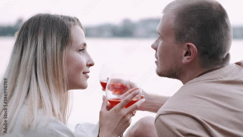 Young man and woan are drinking wine at the lakeside. The couple is enjoying time together on a picnic. Young man and woan are drinking wine at the lakeside. The couple is enjoying time together on a picnic.
