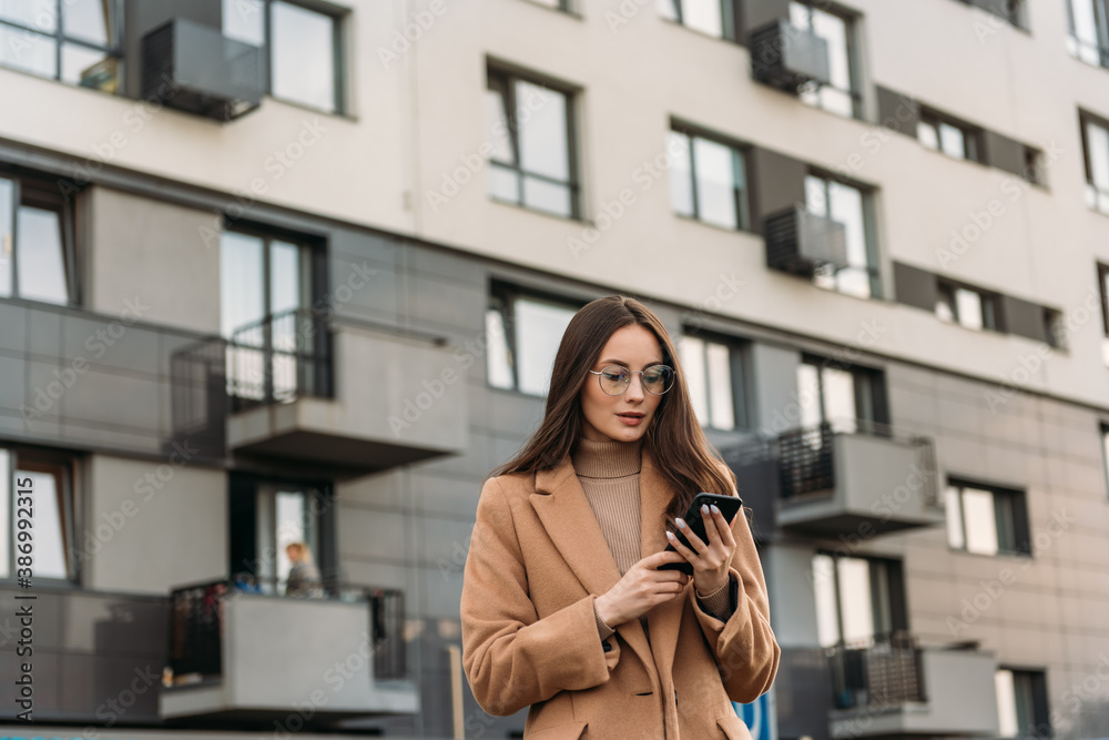 Fototapeta premium Smiling curly businesswoman wearing trendy sunglasses walks down the central city street and uses her phone. Businesswoman holding cell phone.
