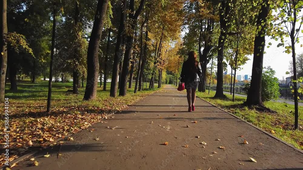Rear view A red-haired woman in red boots and a red bag walks along the alley in autumn on a sunny day. Rear view A red-haired woman in red boots and a red bag walks along the alley in autumn on a sunny day.