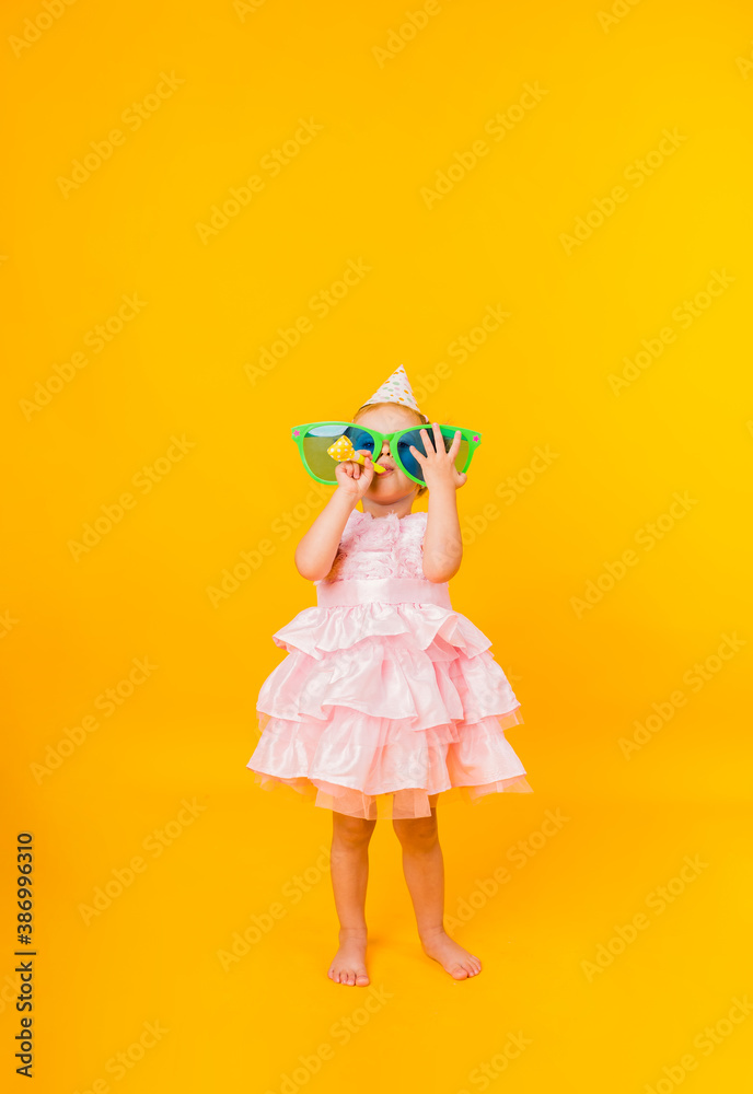 a cheerful little girl in a pink puffy dress holds big funny glasses in her hands and blows a whistle on a yellow background