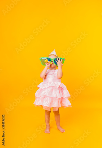 a cheerful little girl in a pink puffy dress holds big funny glasses in her hands and blows a whistle on a yellow background