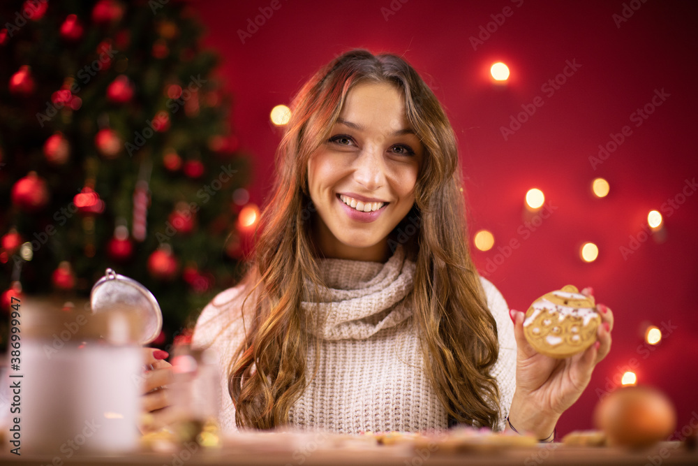 Portrait of Pretty smiling brunette girl decorating freshly baked Christmas cookies on blurred background of Christmas tree and red Xmas decoration. handmade Mindful Gifting. Portrait of Pretty smiling brunette girl decorating freshly baked Christmas cookies on blurred background of Christmas tree and red Xmas decoration. handmade Mindful Gifting.
