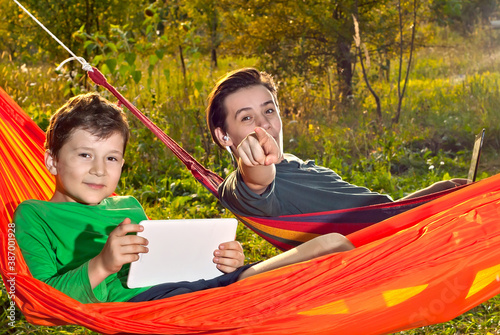 Two boys in orange hammocks on a green background. Children play with laptop and tablet. The concept of online learning in nature. © Ivan
