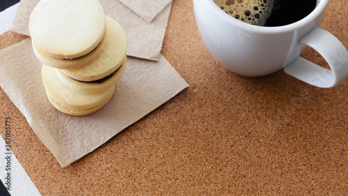 Primer plano desde un ángulo cenital de una taza de cerámica blanca de café negro caliente junto a tres pequeños alfajores rellenos de dulce leche horneados en casa y bajo iluminación natural. 