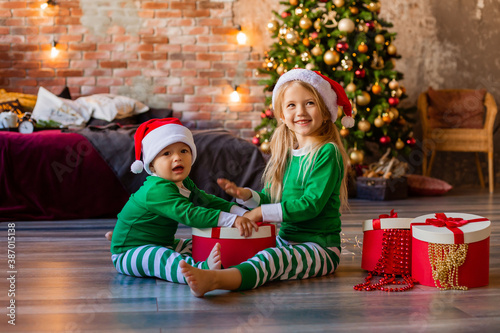 Quadro em tela Two young children in pajamas and Santa hats unpack Christmas presents in the ro