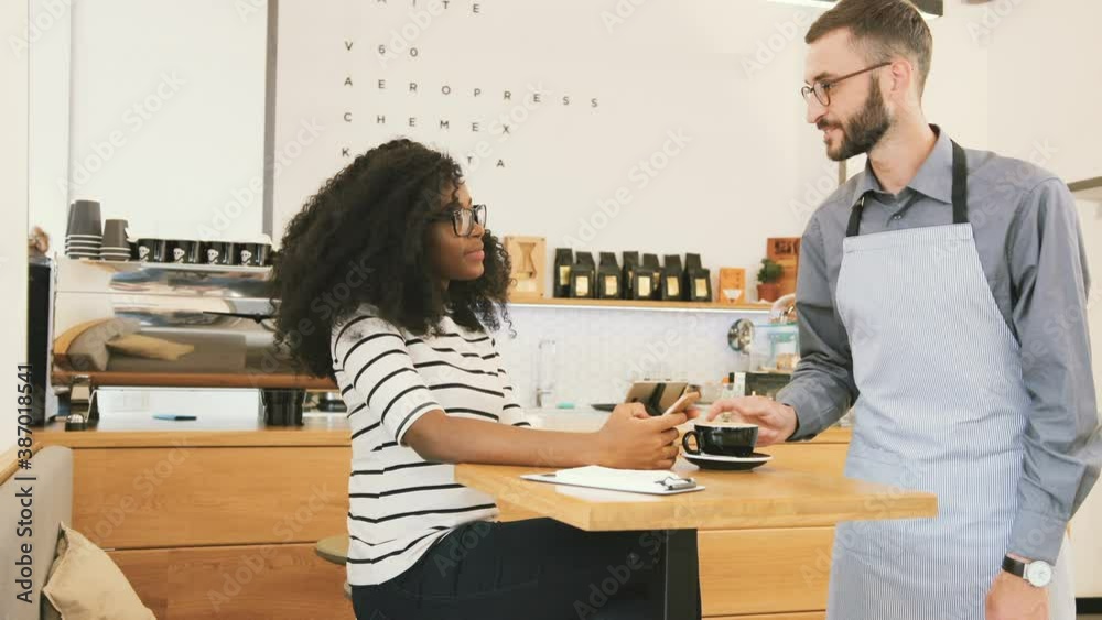 Beautiful African American girl with glasses and black curly hair sitting in cafe and looks into the phone. Young Caucasian waiter brings cup of coffee to client. Beautiful African American girl with glasses and black curly hair sitting in cafe and looks into the phone. Young Caucasian waiter brings cup of coffee to client.