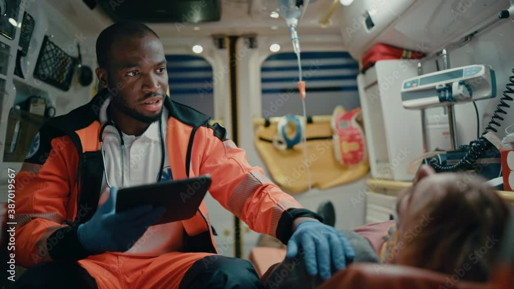 Black EMS Professional Paramedic Using Tablet Computer to Fill a Questionnaire for the Injured Patient on the Way to Hospital. Emergency Care Assistant Comforting the Patient in an Ambulance.