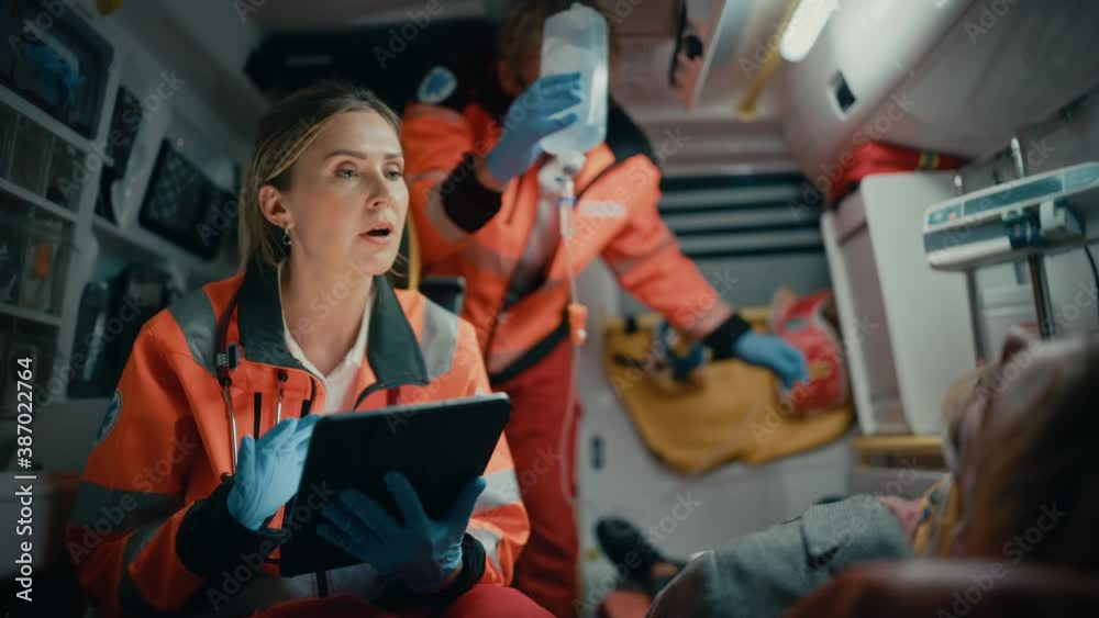 Female EMS Professional Paramedic Using Tablet Computer to Fill a Questionnaire for the Injured Patient on the Way to Hospital. Emergency Care Assistant Comforting the Patient in an Ambulance.