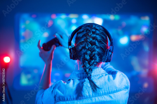 View from back of beautiful gamer woman playing video game at home in front of big screen with joystick and headphone. Colorful neon led. © Anton